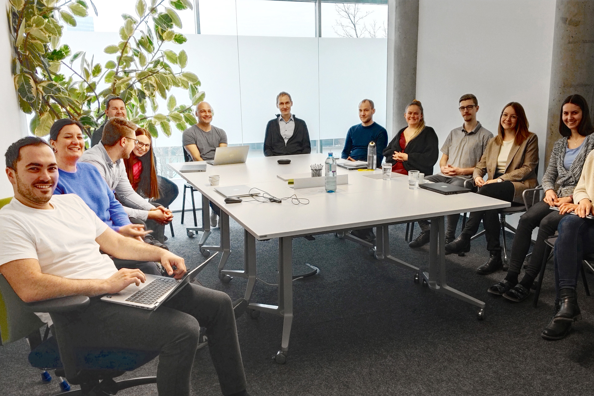 Das Digitrans-Team in einem modernen Büro, versammelt um einen Konferenztisch zu einem kollaborativen Meeting mit Laptops und Notizbüchern. Digitrans team in a modern office setting, gathered around a conference table.