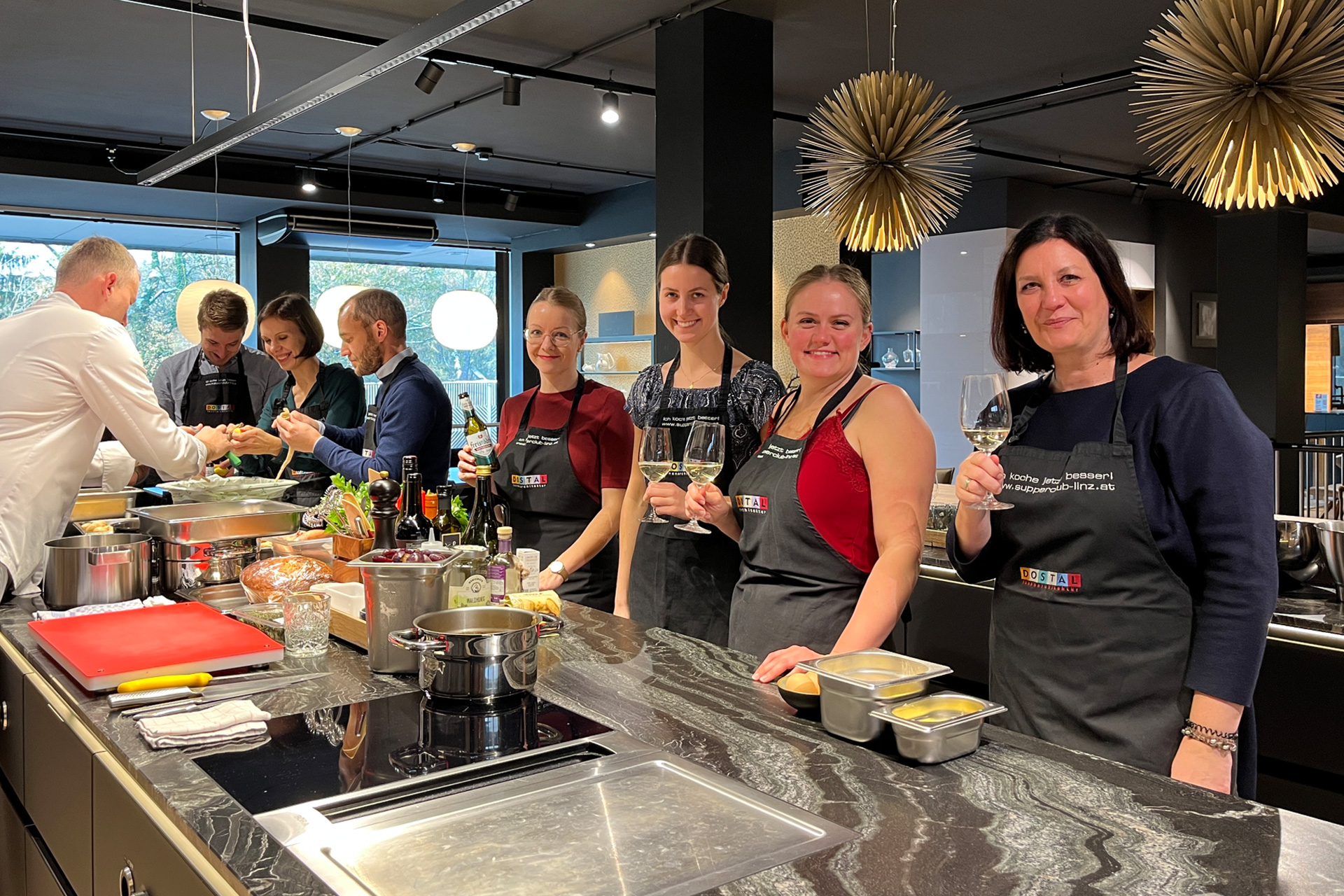 Digitrans-Mitglieder genießen ein gemeinsames Kochevent in einer modernen Küche, tragen gebrandete Schürzen und teilen Lächeln bei Essen und Wein. Digitrans members enjoying a group cooking event in a modern kitchen, wearing branded aprons and sharing smiles over food and wine.