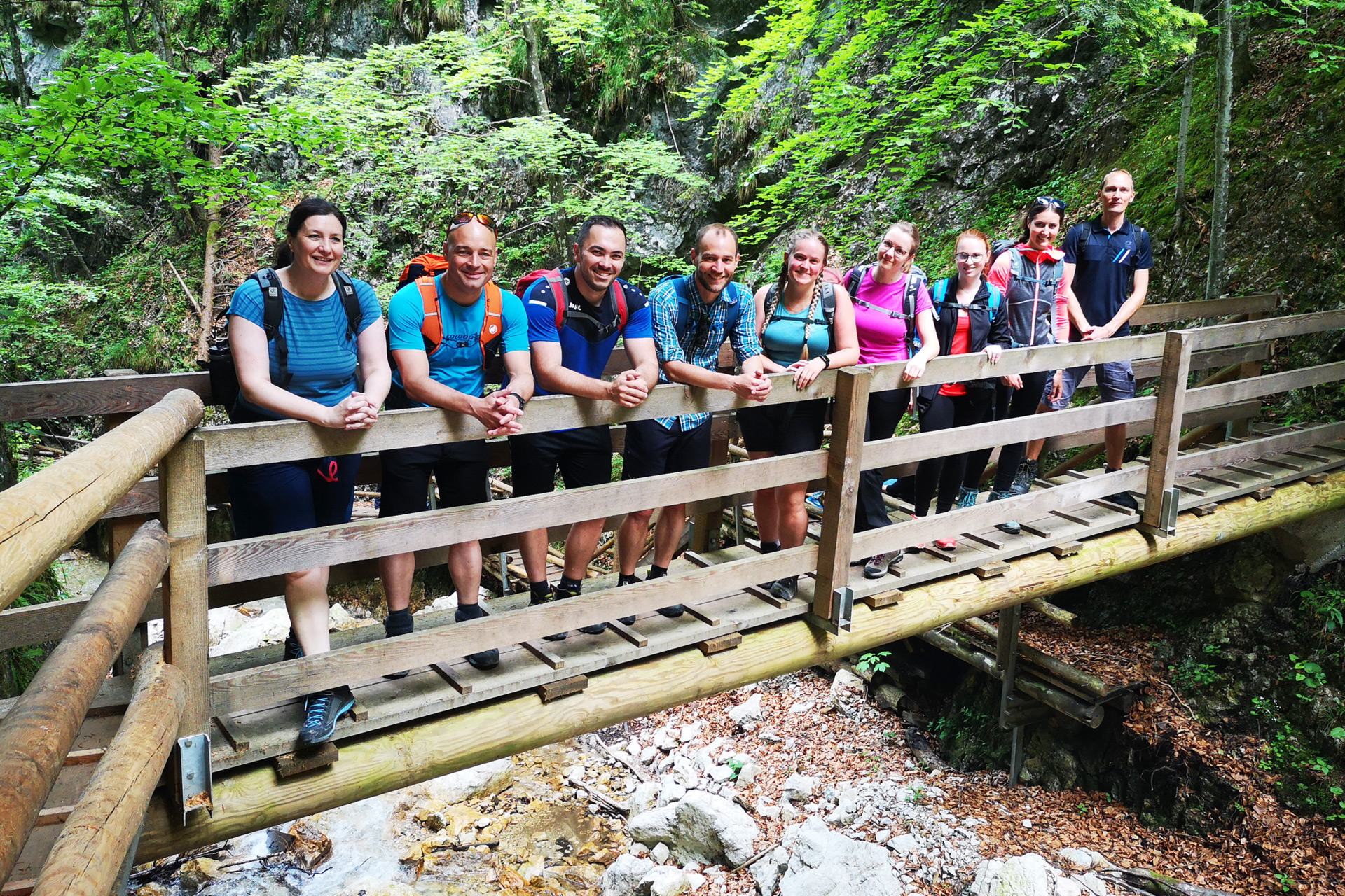 Digitrans-Mitglieder posieren auf einer Holzbrücke während einer Firmenausflug-Wanderung, umgeben von dichtem Wald und einem felsigen Bach, gekleidet in legerer Outdoor-Bekleidung und gemeinsam lächelnd. Digitrans members posing on a wooden bridge during a Firmenausflug hike.