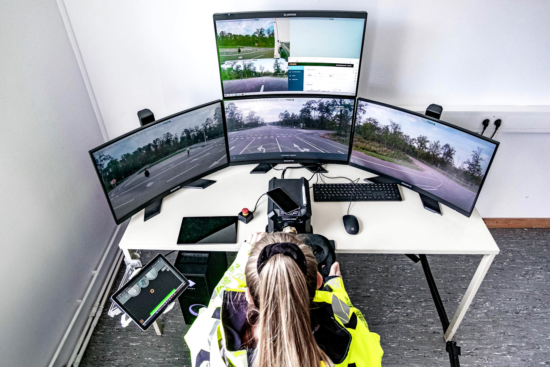 Digitrans-Teleoperations-Setup für automatisiertes Fahren, mit einer Person in einer Warnjacke, die einen Multi-Screen-Fahrsimulator mit Lenkradsteuerung und Datenschnittstellen bedient. Digitrans teleoperation setup for automated driving.