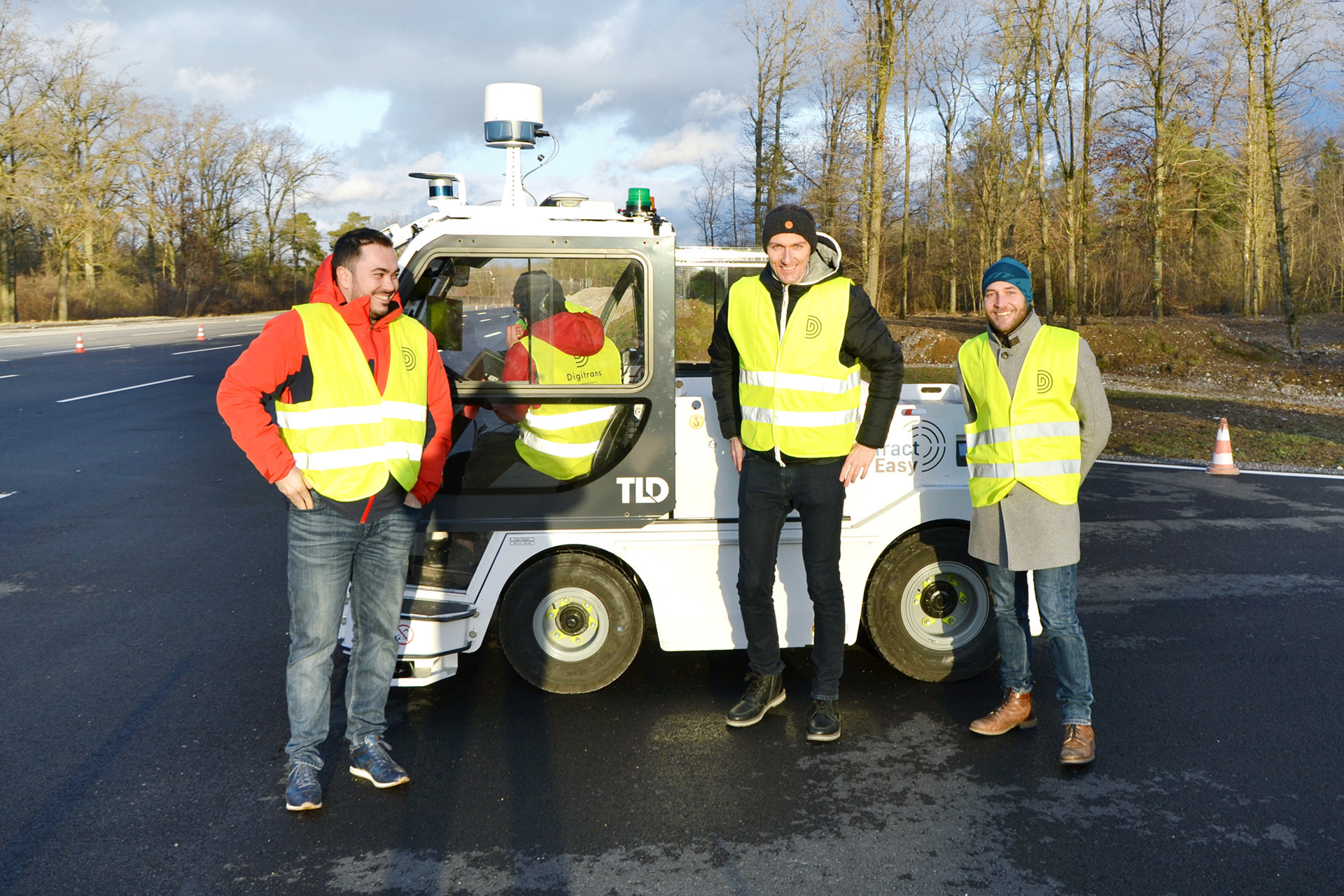 Das Digitrans-Teststreckenteam steht während eines Feldtests um ein autonomes Fahrzeug, trägt Sicherheitswesten in einer kontrollierten Außenumgebung mit Verkehrskegeln und von Wald umgeben. The Digitrans test track team stands around an autonomous vehicle during a field test.